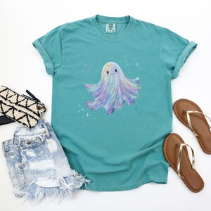May include: Teal-colored t-shirt featuring a whimsical, pastel-colored ghost illustration with a smiling face and sparkling accents. The shirt is paired with denim shorts, a patterned clutch, and brown sandals.