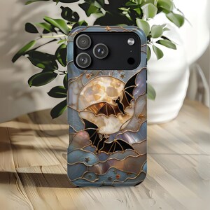 Pu&ograve; includere: Una cover per telefono con un design in stile vetrata che raffigura una grande luna color crema, due pipistrelli neri in volo e piccole stelle dorate. Lo sfondo &egrave; composto da segmenti blu e grigi iridescenti delineati da linee dorate.