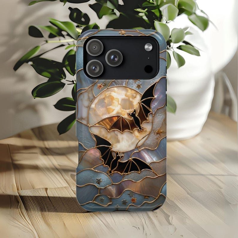 Pu&ograve; includere: Una cover per telefono con un design in stile vetrata che raffigura una grande luna color crema, due pipistrelli neri in volo e piccole stelle dorate. Lo sfondo &egrave; composto da segmenti blu e grigi iridescenti delineati da linee dorate.