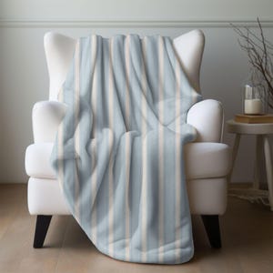 May include: A soft, striped blanket in shades of light blue and cream is draped over a white armchair. The blanket features vertical stripes and a plush texture, creating a cozy and inviting aesthetic. The chair has black legs and is set against a neutral background.