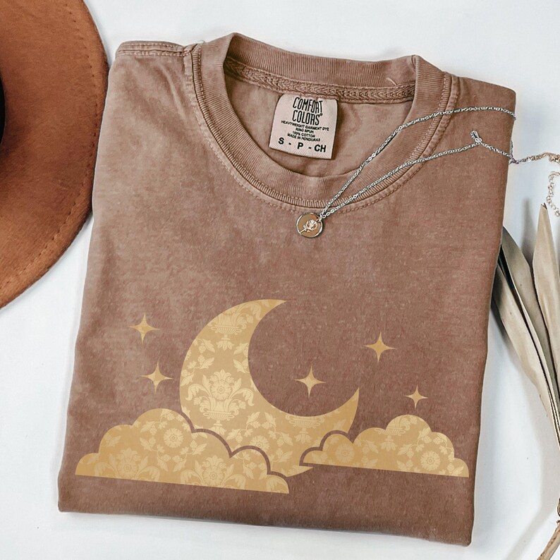 May include: A brown Comfort Colors t-shirt featuring a gold crescent moon and star design. The moon and clouds have a floral pattern. A silver necklace with a small pendant is draped across the shirt.