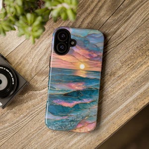 May include: A smartphone case featuring a sunset over the ocean design. The case has a colorful abstract design with shades of blue, pink, and gold. The case is lying on a wooden surface.