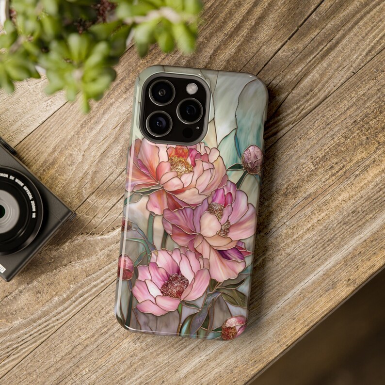 May include: A phone case with a stained glass design featuring pink and white peonies. The case has a cutout for the phone's camera.