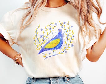 May include: Cream-colored t-shirt featuring a watercolor illustration of a blue and yellow bird perched on a branch, surrounded by a wreath of yellow leaves and blue flowers. The shirt is made of soft material.