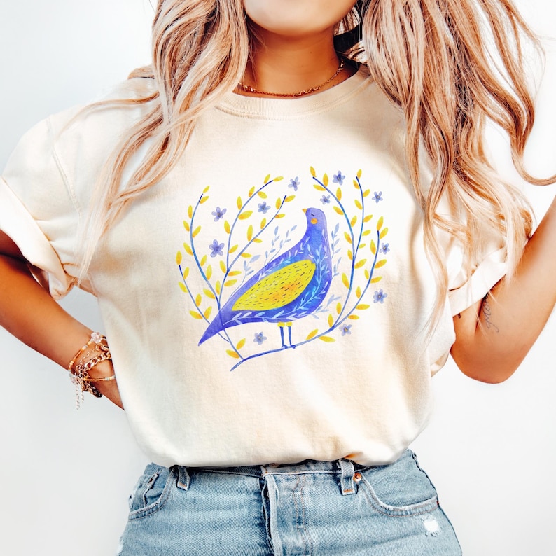 Puede incluir: Camiseta color crema con una ilustraci&oacute;n en acuarela de un p&aacute;jaro azul y amarillo posado en una rama, rodeado de una corona de hojas amarillas y flores azules. La camiseta es de material suave.