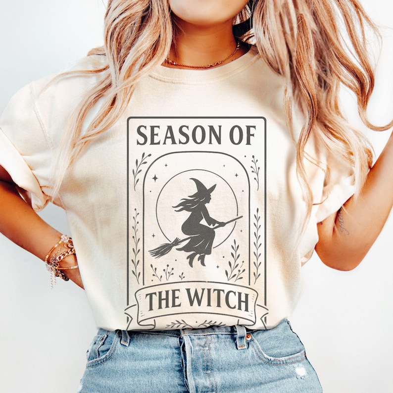 May include: Cream-colored sweatshirt featuring a framed graphic with the text "SEASON OF THE WITCH." The graphic depicts a witch on a broomstick in front of a full moon. The design is in dark gray, with decorative floral elements.