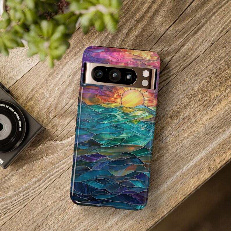 Pu&ograve; includere: Una custodia per telefono colorata con un designo a vetri colorati di un tramonto sull'oceano. La custodia ha un ritaglio per l'obiettivo della fotocamera.