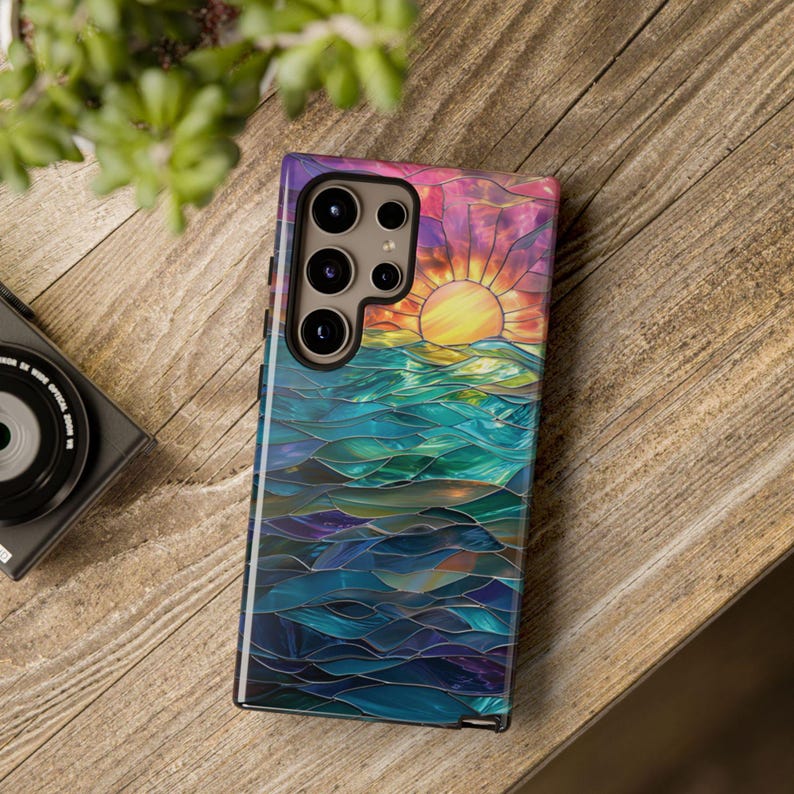 Pu&ograve; includere: Una custodia per telefono colorata con un designo a vetri colorati di un tramonto sull'oceano. La custodia ha un ritaglio per la fotocamera con tre obiettivi.
