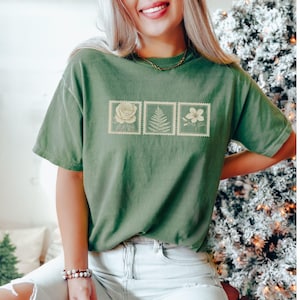 May include: A green t-shirt featuring three postage stamp designs with floral illustrations. The stamps depict a rose, fern, and flower. The shirt is a solid green color and has short sleeves.