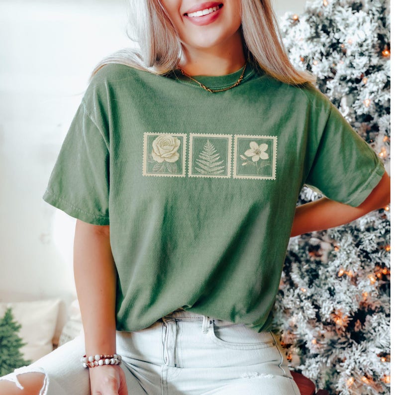 May include: A green t-shirt featuring three postage stamp designs with floral illustrations. The stamps depict a rose, fern, and flower. The shirt is a solid green color and has short sleeves.
