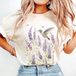 May include: A white t-shirt with a watercolor design of a hummingbird hovering over purple lavender flowers.