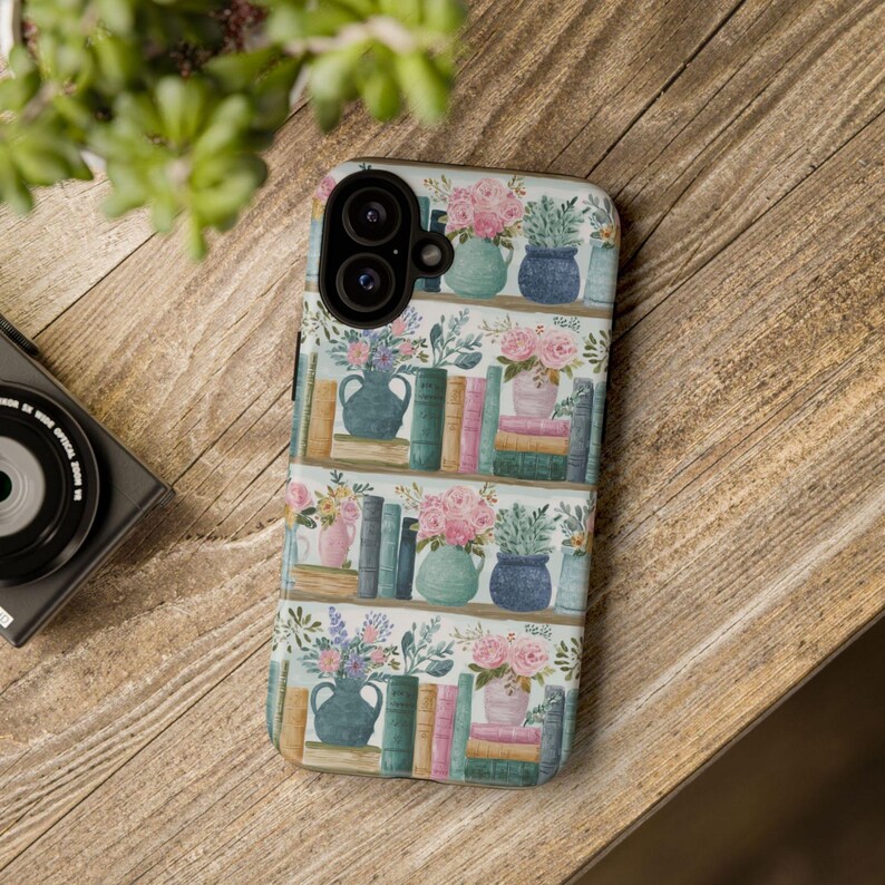 Peut inclure: Une coque de t&eacute;l&eacute;phone avec un motif aquarelle de biblioth&egrave;ques, de vases de fleurs et de piles de livres. Le motif se r&eacute;p&egrave;te sur un fond blanc. La coque est pos&eacute;e sur une surface en bois.