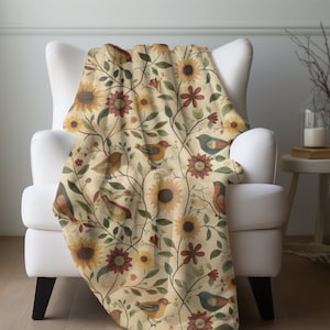 May include: A cream-colored throw blanket with a floral pattern featuring sunflowers, red flowers, and birds. The blanket is draped over a white armchair.