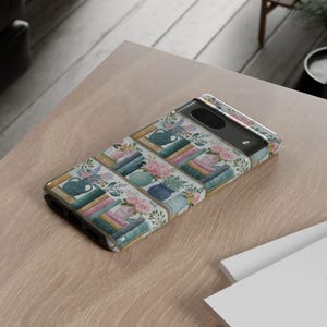Peut inclure: Une coque de t&eacute;l&eacute;phone avec un motif aquarelle de livres, de vases et de fleurs. La coque a un fond blanc avec des illustrations color&eacute;es de livres empil&eacute;s, de vases bleus et d'arrangements floraux roses et violets. La coque est sur une surface marron clair.