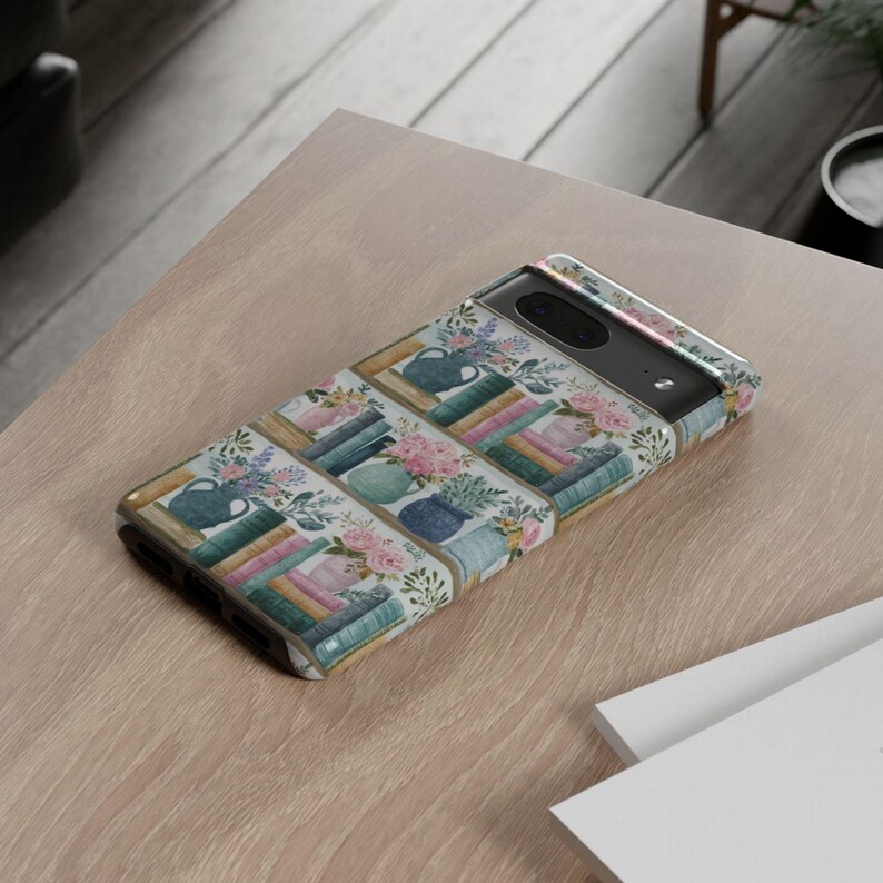 Peut inclure: Une coque de t&eacute;l&eacute;phone avec un motif aquarelle de livres, de vases et de fleurs. La coque a un fond blanc avec des illustrations color&eacute;es de livres empil&eacute;s, de vases bleus et d'arrangements floraux roses et violets. La coque est sur une surface marron clair.