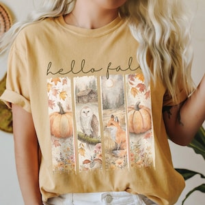 May include: Mustard-colored t-shirt with a fall-themed graphic. The graphic features pumpkins, an owl, a fox, and a cabin, with the words "hello fall" in script. The design has a watercolor style.