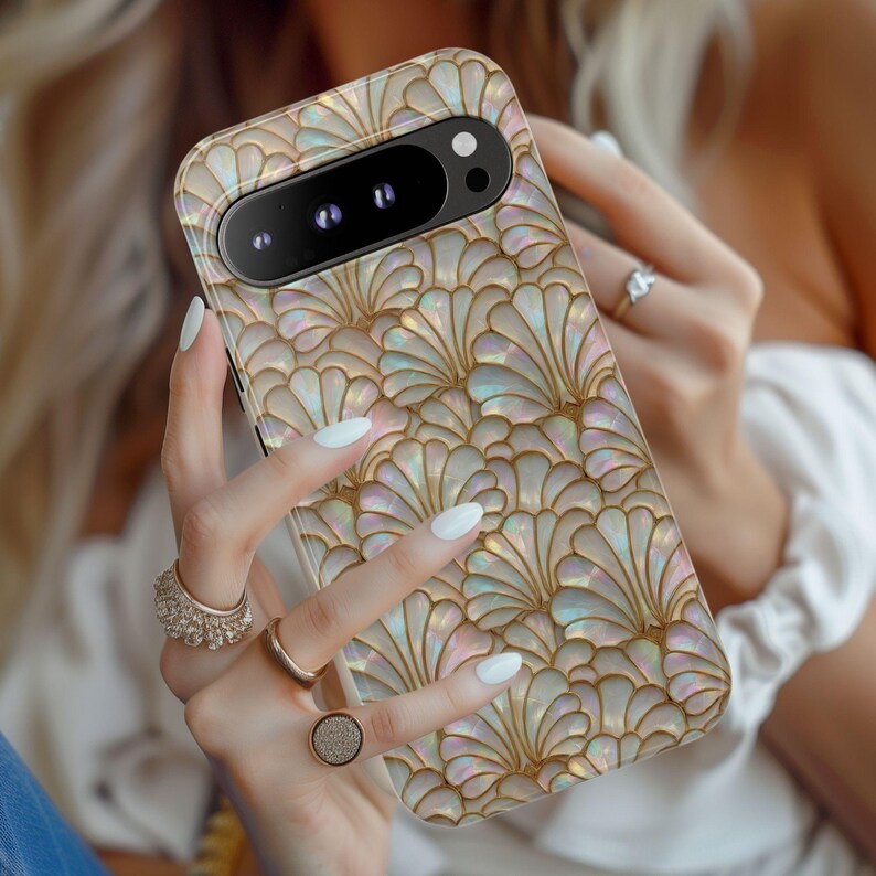 May include: A phone case with a pearlescent, fan-shaped pattern in gold and iridescent hues. The case has a camera cutout and is held by a person with white nail polish and gold rings. The case is designed for a smartphone.