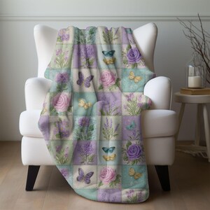 May include: A patchwork throw blanket draped over a white armchair. The blanket features a repeating pattern of squares with floral and butterfly designs in pastel colors, including lavender, pink, and teal. The blanket is made of a soft material.