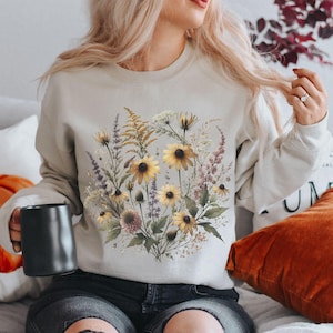 May include: A beige sweatshirt featuring a floral design with yellow and brown flowers, green leaves, and purple accents. The sweatshirt is a neutral color and has a crew neck. A black mug is held in the left hand.