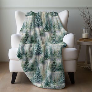 May include: A cozy throw blanket draped over a white armchair. The blanket features a watercolor forest scene with evergreen trees in shades of green and blue against a soft, light background. The blanket adds a touch of nature-inspired comfort.
