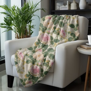 May include: A soft, cream-colored throw blanket with a floral pattern of pink and yellow roses and green leaves. The blanket is draped over a white armchair.