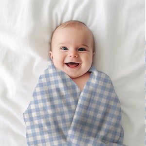 May include: A baby wrapped in a blue and white gingham swaddle blanket, smiling at the camera.