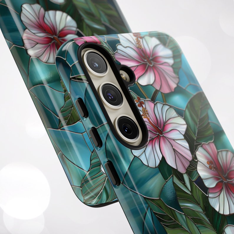 May include: A teal blue phone case with a stained glass design featuring pink and white hibiscus flowers. The case has a raised camera bump with three lenses.