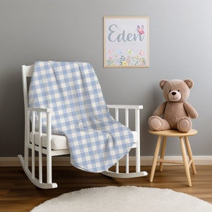 May include: A white rocking chair with a blue and white checkered blanket draped over it. A teddy bear sits on a wooden stool next to the chair. A framed picture with the name "Eden" and butterflies hangs on the wall.