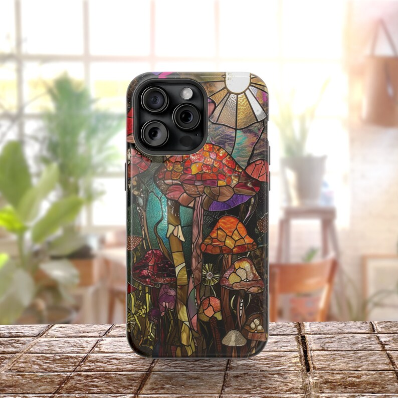 May include: A phone case with a stained glass design featuring colorful mushrooms and a sun. The case is black and has a raised camera bump.