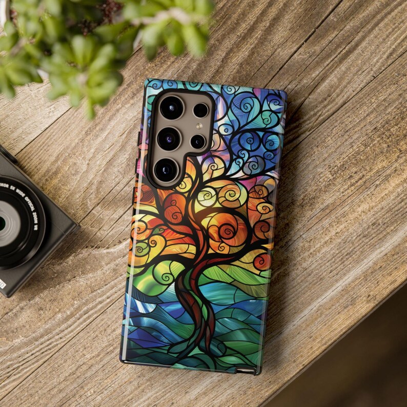 May include: A colorful phone case with a stained glass design of a tree with swirling branches. The case is designed for a phone with a triple lens camera.