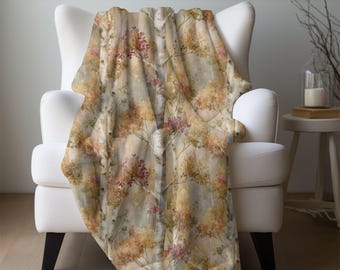 Può includere: Una morbida coperta in peluche drappeggiata su una poltrona bianca. La coperta presenta un design ispirato alla natura con tronchi e rami di betulla, adornati da foglie dai colori autunnali nelle tonalità dell'oro, dell'arancio e del bordeaux. Un elemento d'arredo per la casa.