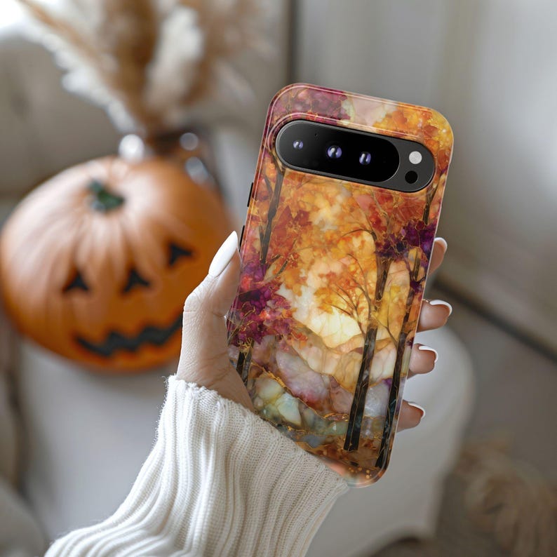 May include: A phone case featuring an autumn landscape with trees, colorful foliage, and a mountain range. The case has a clear, glossy finish and is held in a hand. The background includes a carved pumpkin and a vase with dried flowers.