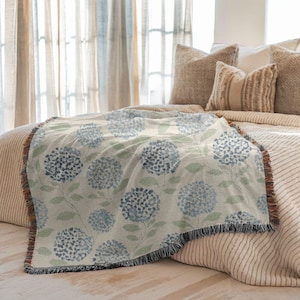 May include: A woven throw blanket featuring a floral hydrangea pattern in shades of blue and green on a cream background. The blanket has a fringed edge and is draped over a bed with neutral-toned pillows.