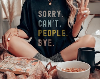May include: A woman wearing a black t-shirt with the text "SORRY. CAN'T. PEOPLE. BYE." in colorful letters. She is sitting on a bed with a blanket and a bowl of cereal.