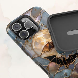 Pu&ograve; includere: Una custodia per telefono con un design in stile vetrata, caratterizzata da segmenti iridescenti blu, bianchi e ambrati delineati in oro. Il motivo include sagome di pipistrelli neri e piccole stelle dorate. La custodia ha un interno nero e un foro per la fotocamera.