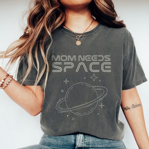 May include: A gray t-shirt with the text "MOM NEEDS SPACE" in white outlined letters. The text is above a graphic of a planet with rings and stars.