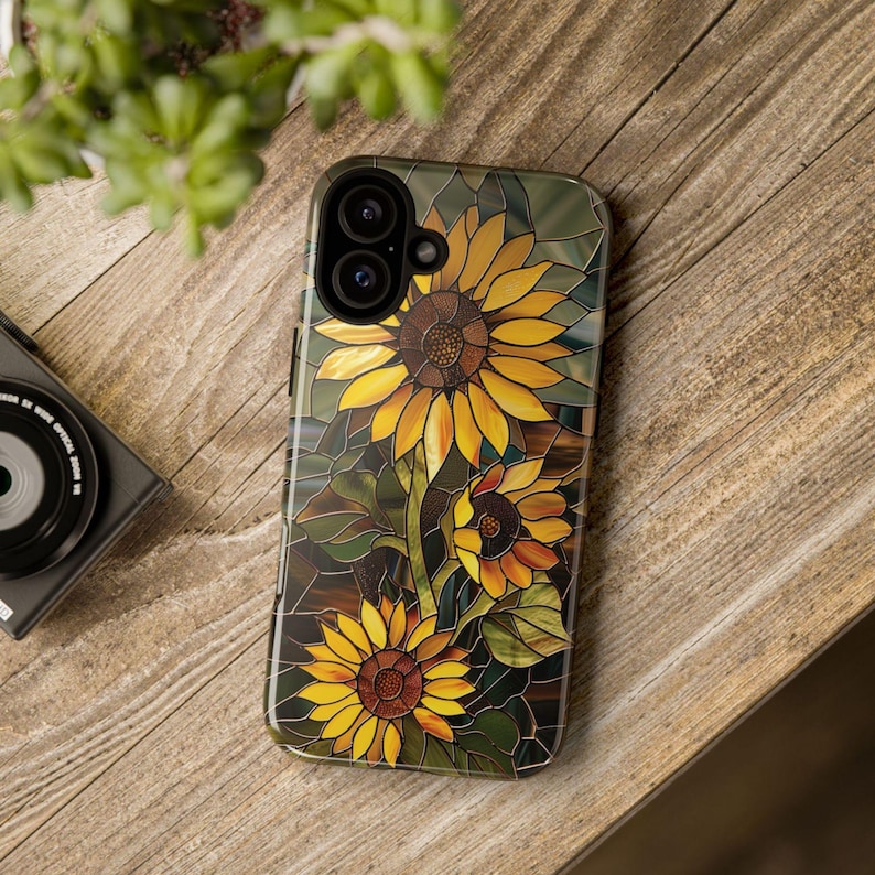 May include: A phone case with a stained glass design featuring three sunflowers in shades of yellow, orange, and brown. The case has a black camera cutout and a clear background.