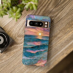 May include: A smartphone case featuring a sunset over the ocean design. The case has a colorful, abstract design with shades of blue, pink, and gold. The phone case is on a wooden surface.