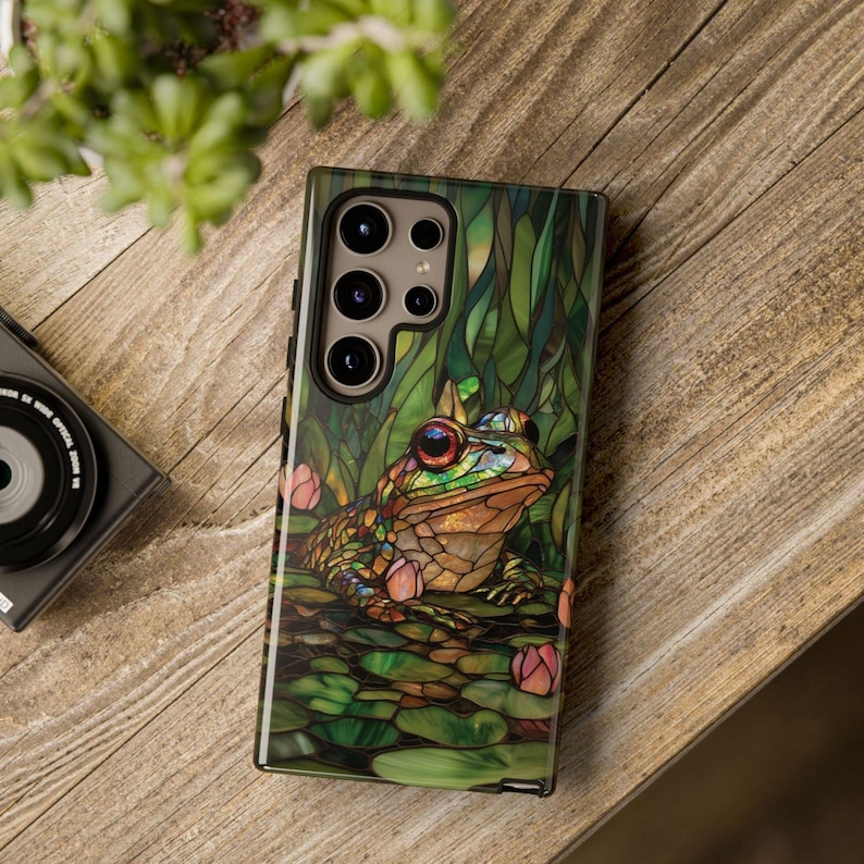 May include: A green, brown, and gold phone case with a stained glass design of a frog sitting on a lily pad. The case has a cutout for the phone's camera.