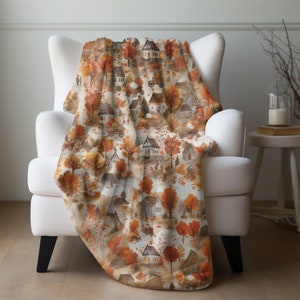May include: A soft, cozy throw blanket featuring a watercolor pattern of autumnal houses, trees, and leaves. The design is repeated throughout the blanket, creating a warm and inviting feel.