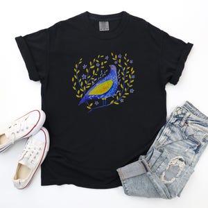 Puede incluir: Camiseta negra con un dise&ntilde;o de p&aacute;jaro colorido. El p&aacute;jaro es azul y amarillo, rodeado por una corona de hojas amarillas y peque&ntilde;as flores azules. La camiseta se combina con vaqueros de mezclilla descoloridos y zapatillas blancas.