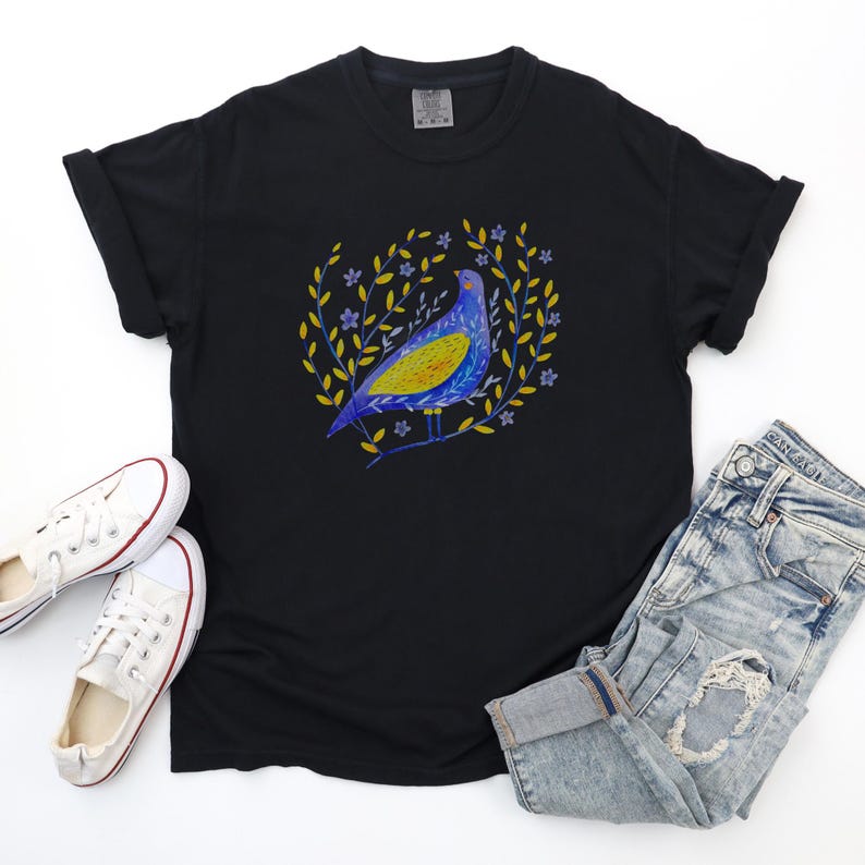 Puede incluir: Camiseta negra con un dise&ntilde;o de p&aacute;jaro colorido. El p&aacute;jaro es azul y amarillo, rodeado por una corona de hojas amarillas y peque&ntilde;as flores azules. La camiseta se combina con vaqueros de mezclilla descoloridos y zapatillas blancas.