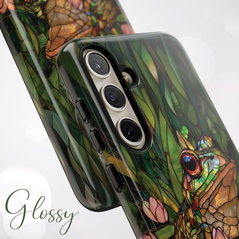 May include: A glossy phone case with a stained glass design featuring a frog, leaves, and flowers. The case has a black camera cutout and a green, brown, and pink color scheme.
