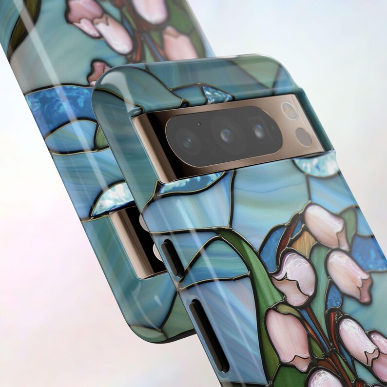 Pu&ograve; includere: Una custodia per telefono blu e verde con un design a vetri colorati di fiori rosa. La custodia ha un rialzo per la fotocamera rialzato con tre obiettivi.