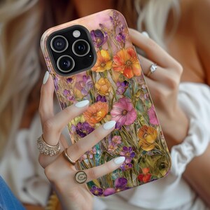 Op de afbeelding: Een telefoonhoesje met een bloemenontwerp in tinten oranje, roze en paars. De hoes heeft een glanzende afwerking en een zwarte cameralensbeschermer. De achtergrond is zachtroze en de algehele esthetiek is artistiek en vrouwelijk.