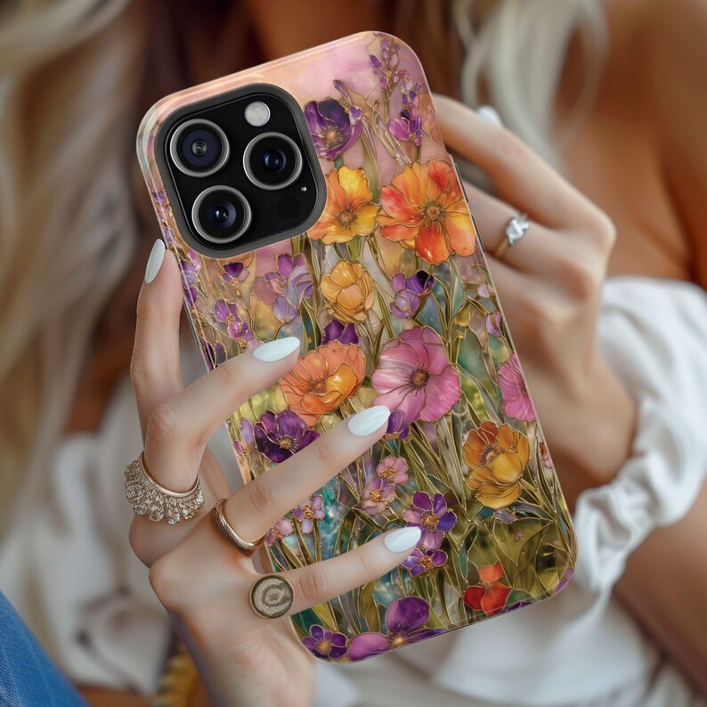 Op de afbeelding: Een telefoonhoesje met een bloemenontwerp in tinten oranje, roze en paars. De hoes heeft een glanzende afwerking en een zwarte cameralensbeschermer. De achtergrond is zachtroze en de algehele esthetiek is artistiek en vrouwelijk.