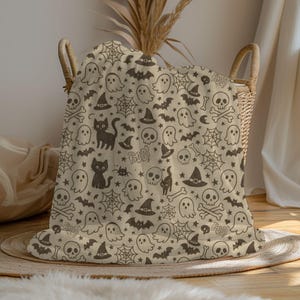 May include: A beige Halloween-themed blanket with a repeating pattern of black cats, ghosts, witch hats, spiderwebs, bats, skulls, and the word "Boo!". The blanket is draped over a wicker basket.