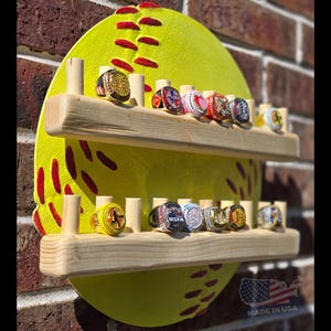 May include: A wooden softball ring holder with two shelves. The holder is painted yellow with red stitching and is mounted on a brick wall. The shelves are filled with championship rings.