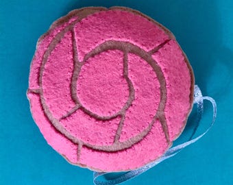 May include: A pink felt ornament shaped like a Mexican sweet bread, called a concha, with a brown felt outline. The ornament has a white ribbon attached for hanging.