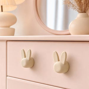 May include: Two light beige drawer knobs shaped like rabbit heads with upright ears. The knobs are mounted on a smooth, pale pink drawer front. Each knob features a rounded face and two distinct, elongated ears extending from the top section.
